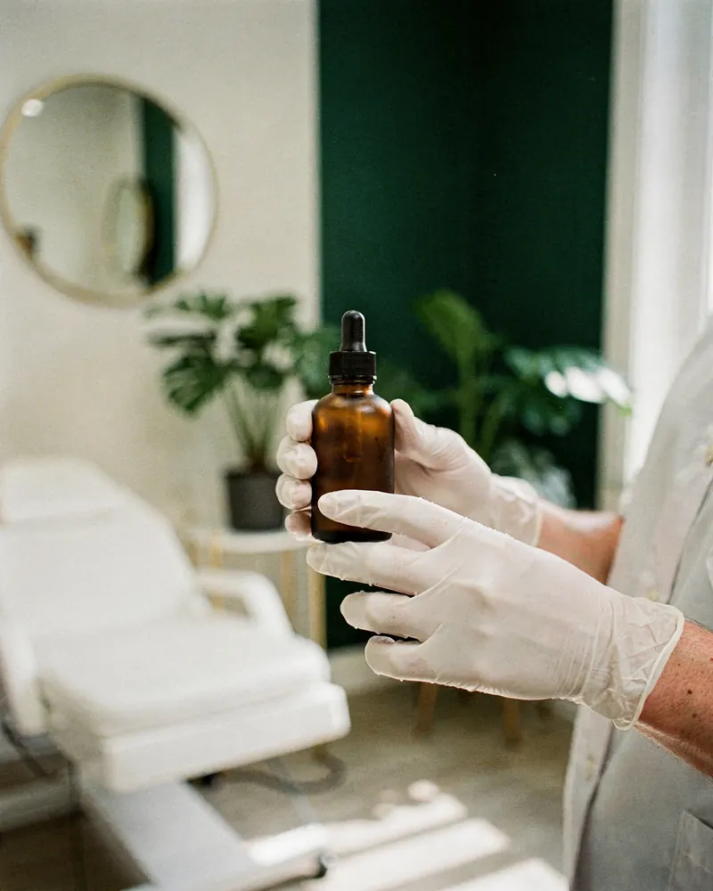 Gloved hands hold an amber serum bottle in a bright treatment room with a procedure chair, round mirror and plants.