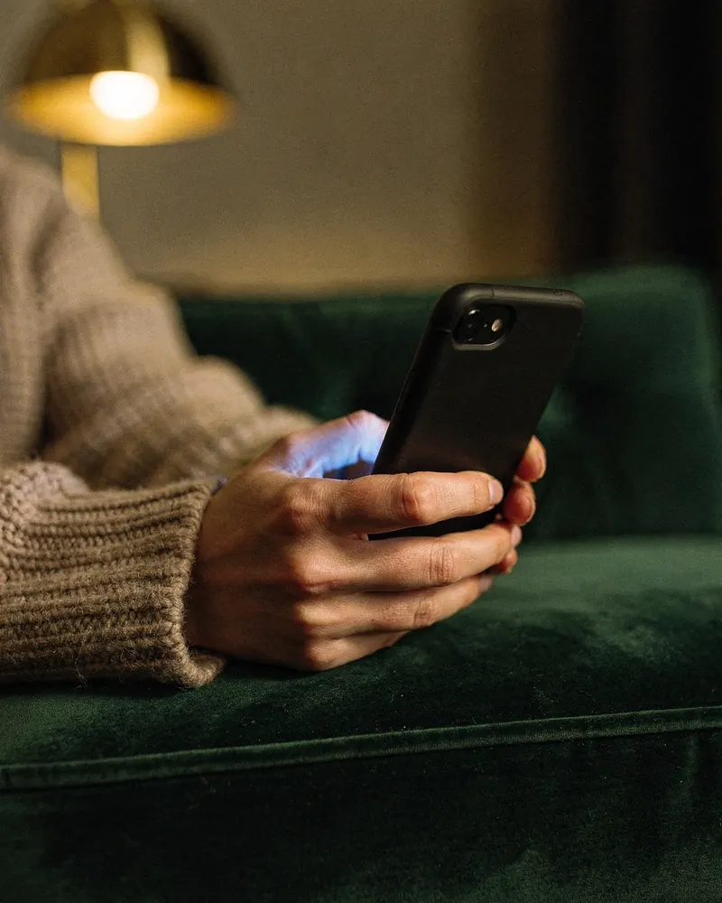 At night on a green velvet sofa, a hand holds a phone whose screen glow lights the fingers; a brass lamp glows behind.
